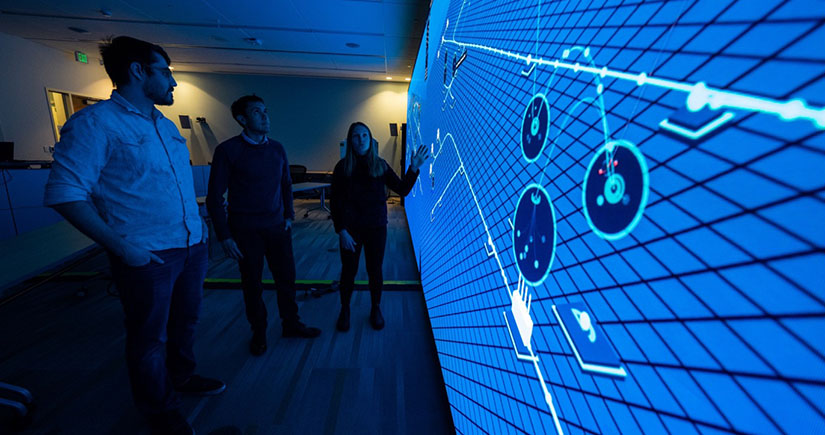 cybersecurity-threat-analysis Three people stand in a dark room looking at a large wall display computer screen.