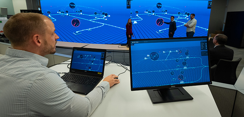 Photo of a researcher on the computer sitting at a desk and two researchers standing in front of a large monitor emulating a real-time cyberattack.