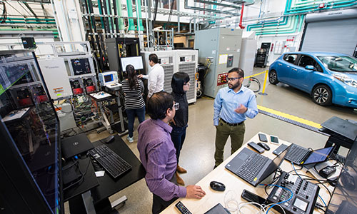 People work together in front of computers on a desk inside laboratory with EV in the background.