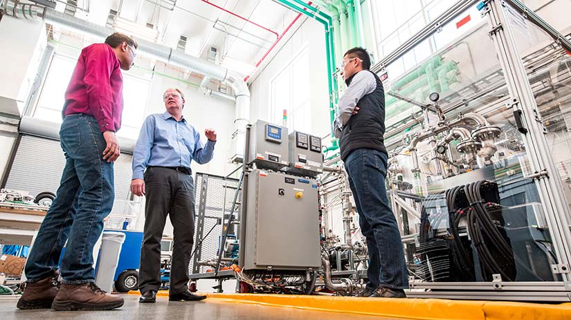 Photo of three researchers talk in front of a hydrogen electrolyzer, one of the unique research capabilities of the ESIF.