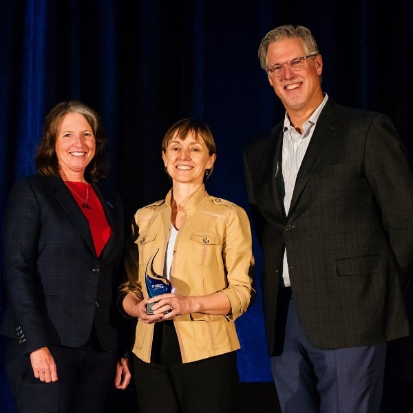 Three people stand in front of a blue curtain in business dress, with the person in the middle holding an award.