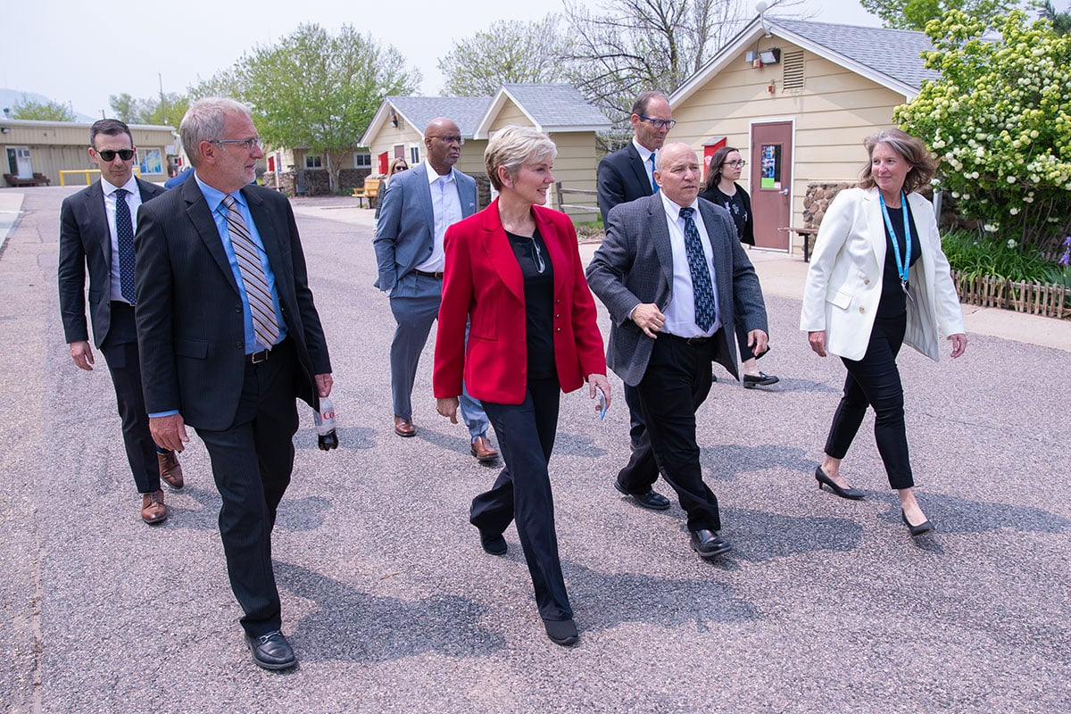 A group of people in business dress walk on a gravel road with alongside buildings.