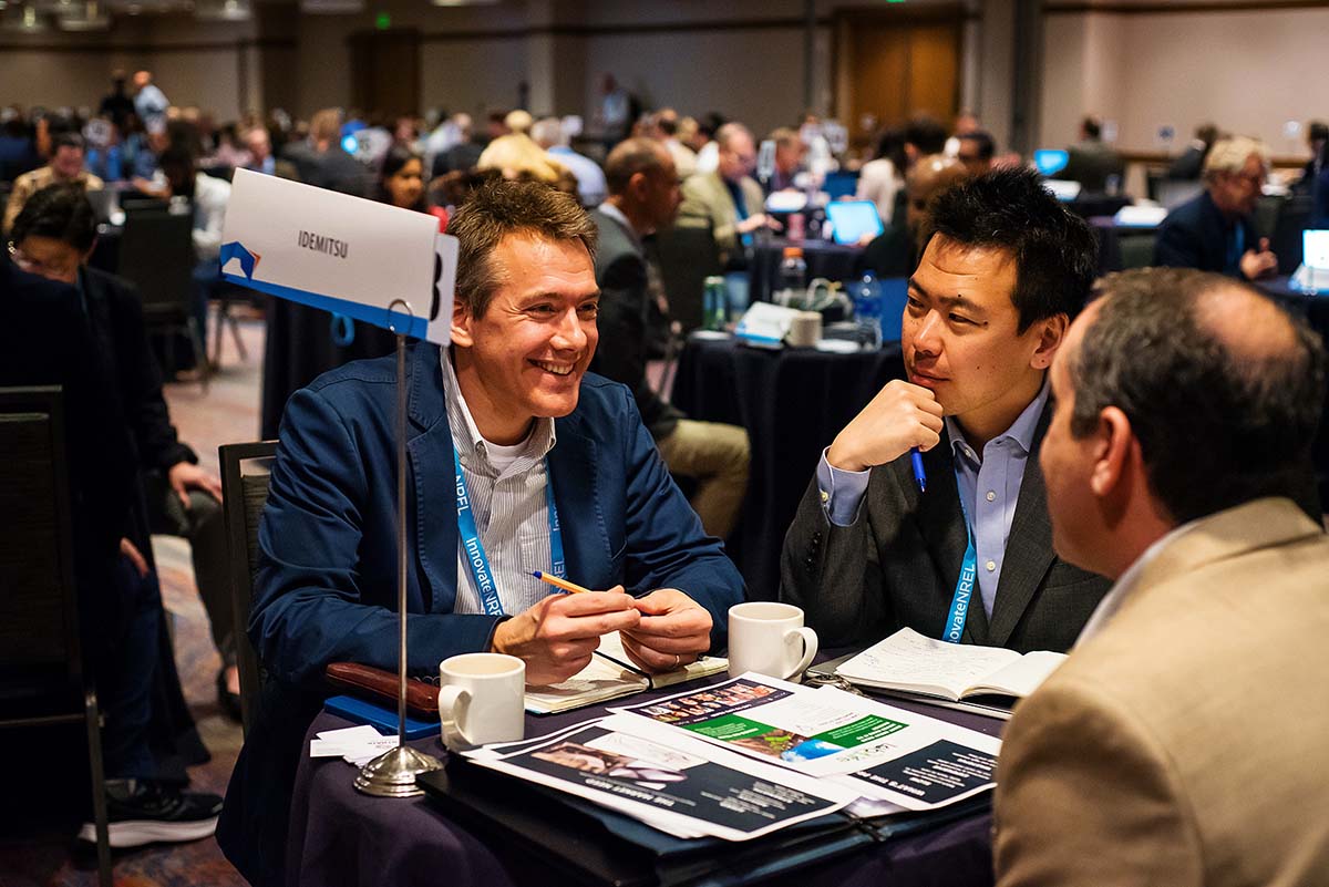 Three people sit at a small table for a meeting in a ballroom with dozens of similarly occupied small tables around them.