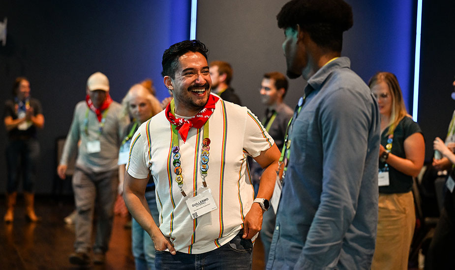 A man in a red bandana smiles and speaks with a man in a blue shirt in a crowded room.