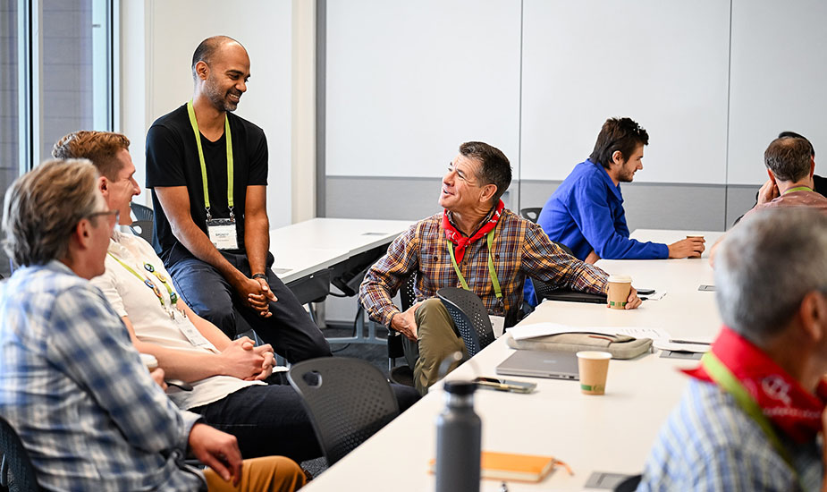 A man in a black shirt speaks with other men who are sitting in a classroom.