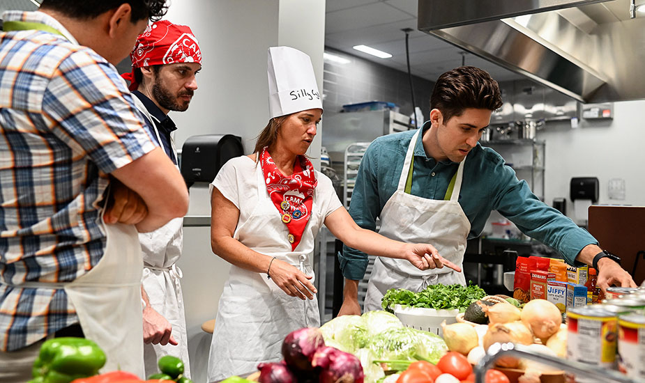 Four people in chef aprons converse with each other while looking at a table of fresh ingredients.