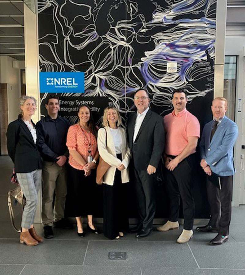 Seven people pose in front of a wall bearing a graphic and the words NREL – Transforming Energy – Energy Systems Integration Facility