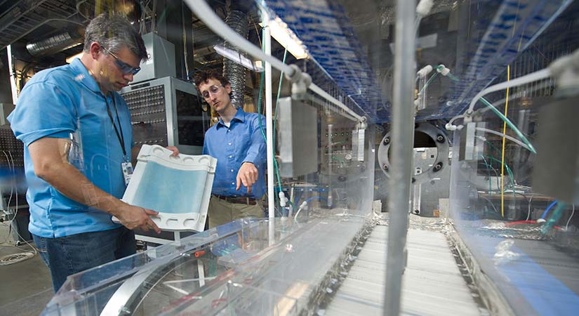 20171 Two researchers communicate while standing on the side of a long piece of machinery.