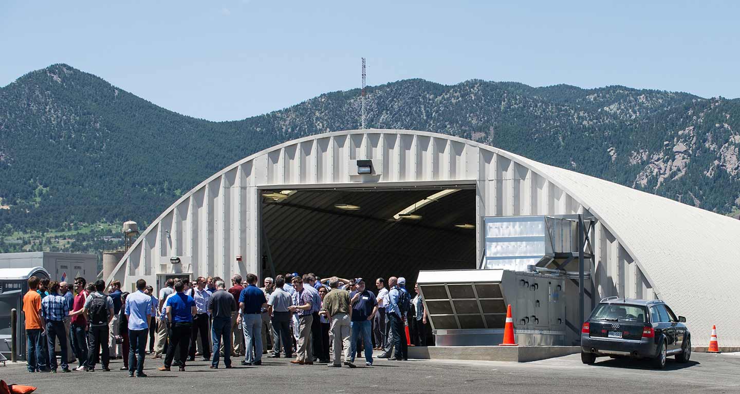 iacmi-hero A large group of people stand outside of a white, half-cylindrical facility with mountains in the background.