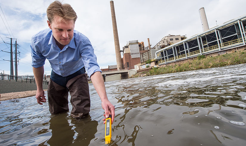 jordan-macknick-33322 A man in a dress shirt standing in front of a power plant gets a temperature reading and water samples from a body of water.