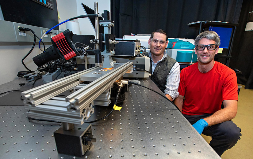 Two men smile at the camera while posing with technical equipment in laboratory setting.
