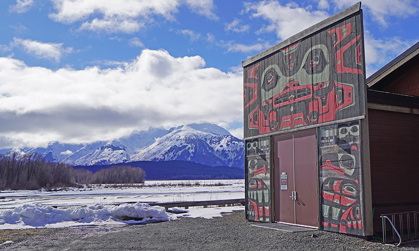 20250507-nrel-partners-with-chilkat-indian-village-to-improve-housing-in-klukwan-alaska-cultural-center A building sits next to a snow-covered river with mountains in the background.