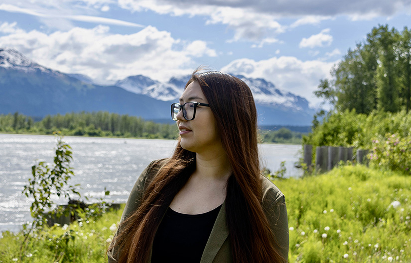 20250507-nrel-partners-with-chilkat-indian-village-to-improve-housing-in-klukwan-alaska-shawna A person with long hair stands in front of a mountainous landscape.
