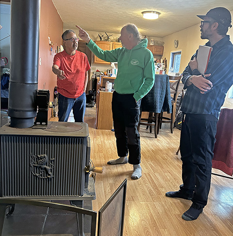 20250507-nrel-partners-with-chilkat-indian-village-to-improve-housing-in-klukwan-alaska-stove Three people stand around a wood-burning stove in a house.
