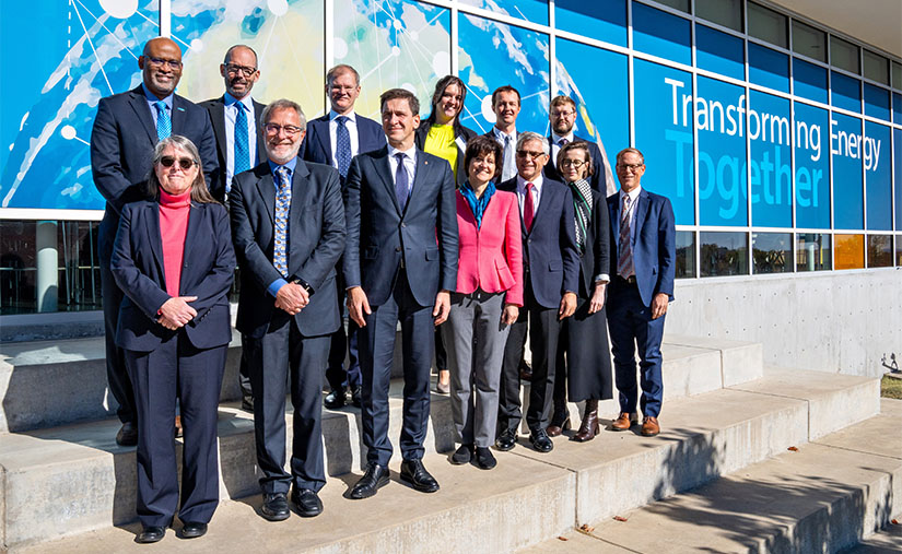 A group of 13 people stand outside in two rows smiling at the camera. Behind them is a background that says
