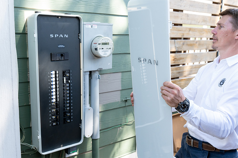 20250416-smart-electrical-panel-startup-spans-nrels-accelerator-programs-outside A man holds the cover of a power panel next to the panel hanging on the outside of a home.
