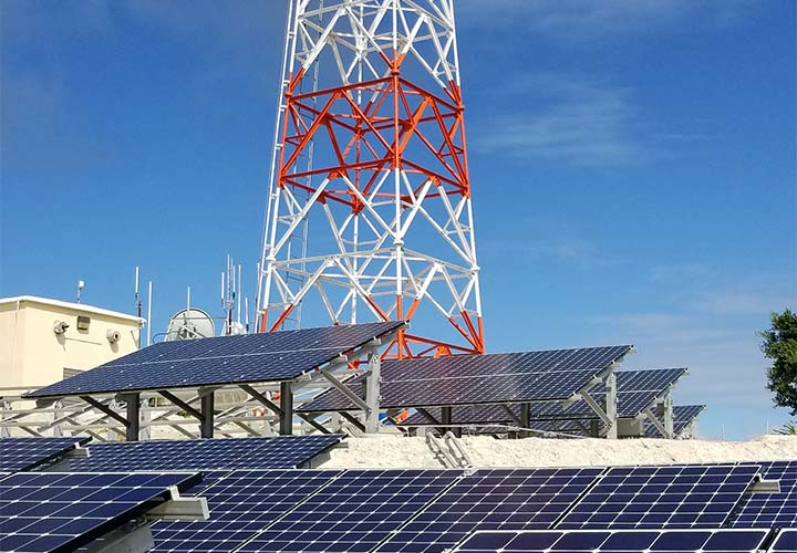 Rows of solar panels with a red and white tower on top of a building behind them.