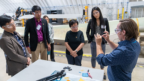 Group of people stand inside laboratory looking at tiny wind turbine components.