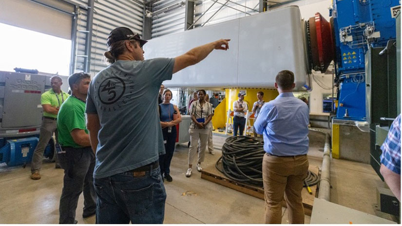 A person in a hat points to a large wind turbine gearbox attached to a dynamometer while a tour group looks on