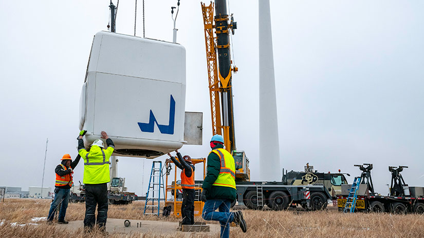 Crews wearing protective gear position a large wind turbine component being lowered by a crane.