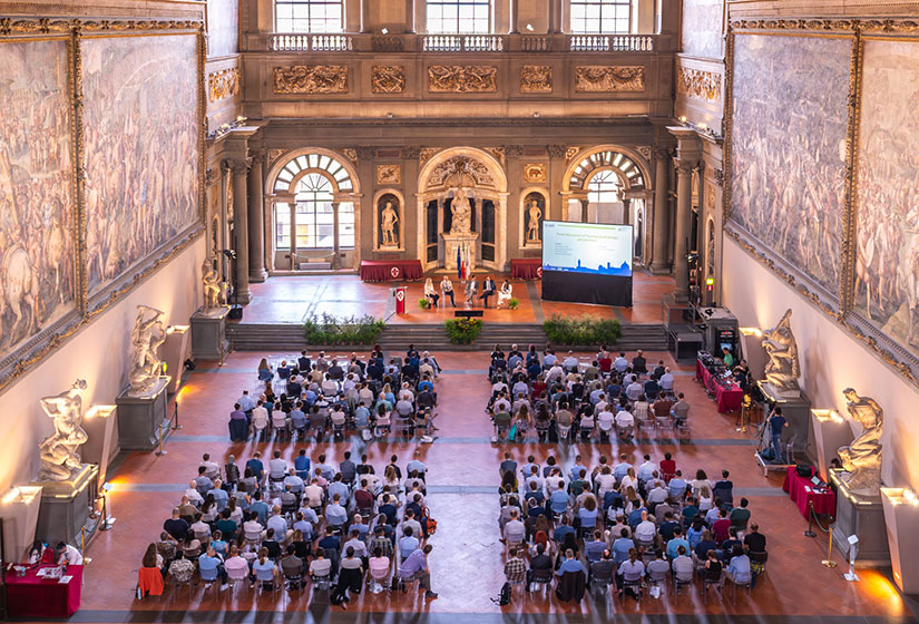 Five people sit on a stage with a screen with about 200 people seated on the floor in front of them. Large paintings and sculptures surround the room