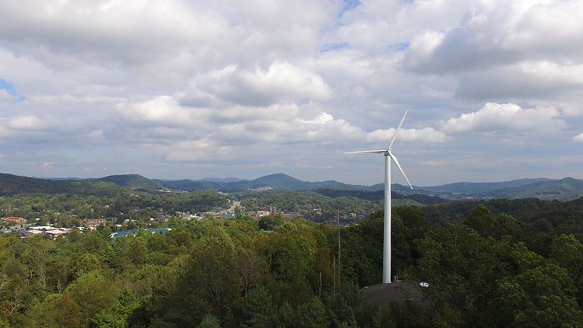 A wind turbine above tree line with mountains in the background