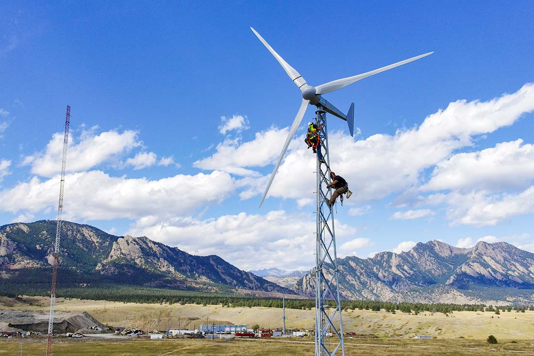 Two workers wearing protective gear work near the top of a small wind turbine with mountains in the background.