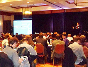 Photo of a seated audience with a presenter at a podium and a projection screen in the background.