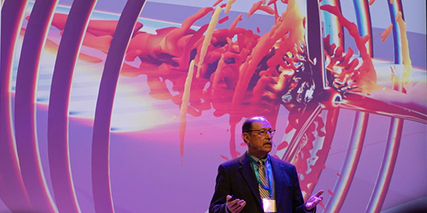 A person in a suit standing in front of a screen displaying a wind turbine model.