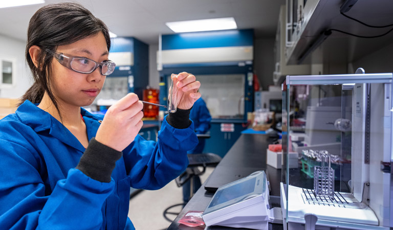 A person wearing protective eyewear and a labcoat holding a vial.