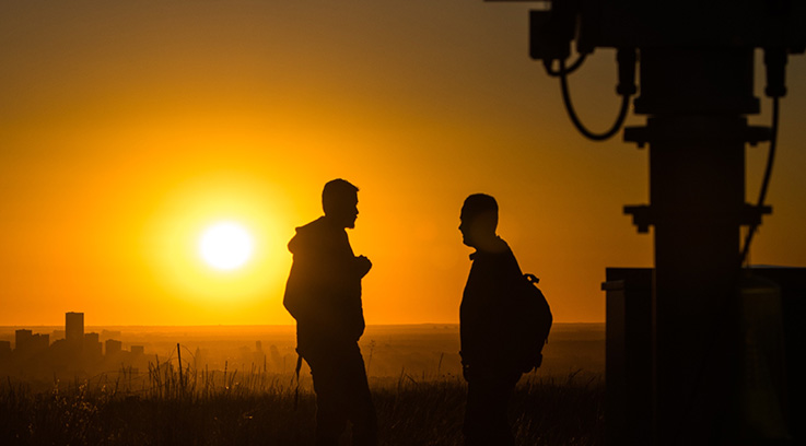 Two people standing in a field with sun beaming behind them.