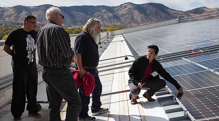 A person kneels near and points at a set of solar panels while three others watch.