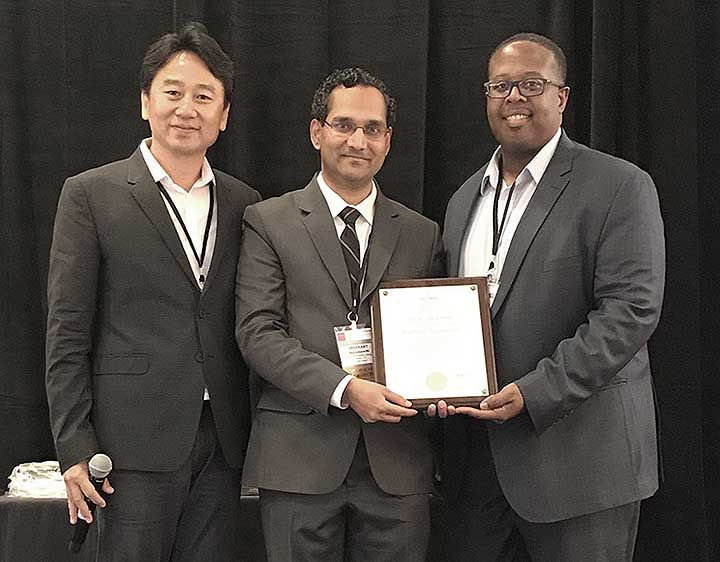 2018-09-photo-06 Three men dressed in suits stand on a stage in front of a black curtain. One man holds a microphone the other two hold an award plaque.