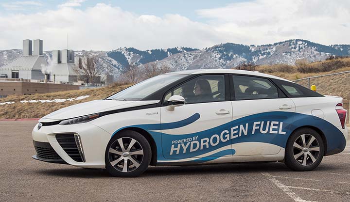 2018-09-photo-10 A female driver sits at the wheel of a hydrogen fuel cell vehicle at NREL with mountains and a building in the background. The vehicle has a decal wrap that reads Powered by Hydrogen Fuel.